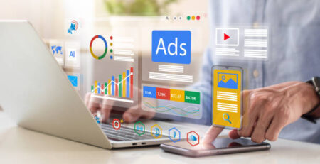 Person interacting with a laptop as holographic marketing dashboards, ads tiles, and charts float above the screen and a smartphone nearby.