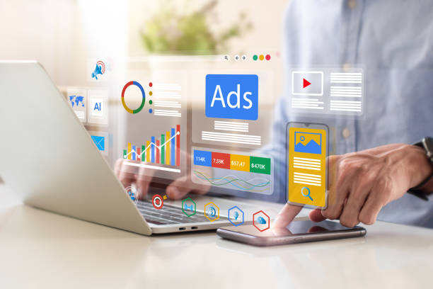 Person interacting with a laptop as holographic marketing dashboards, ads tiles, and charts float above the screen and a smartphone nearby.